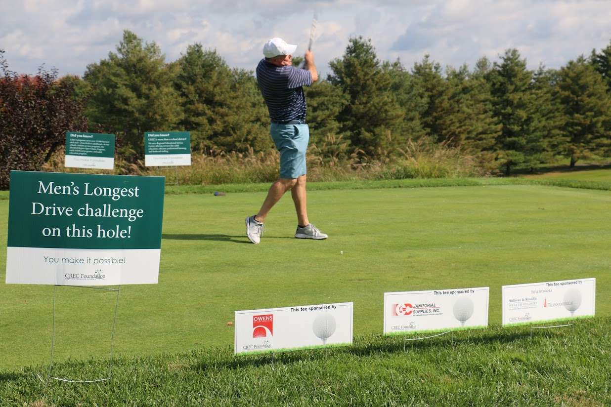A golfer tees off in front of a Men's Longest Drive Challenge sign, as well as other sponsorship tee signs